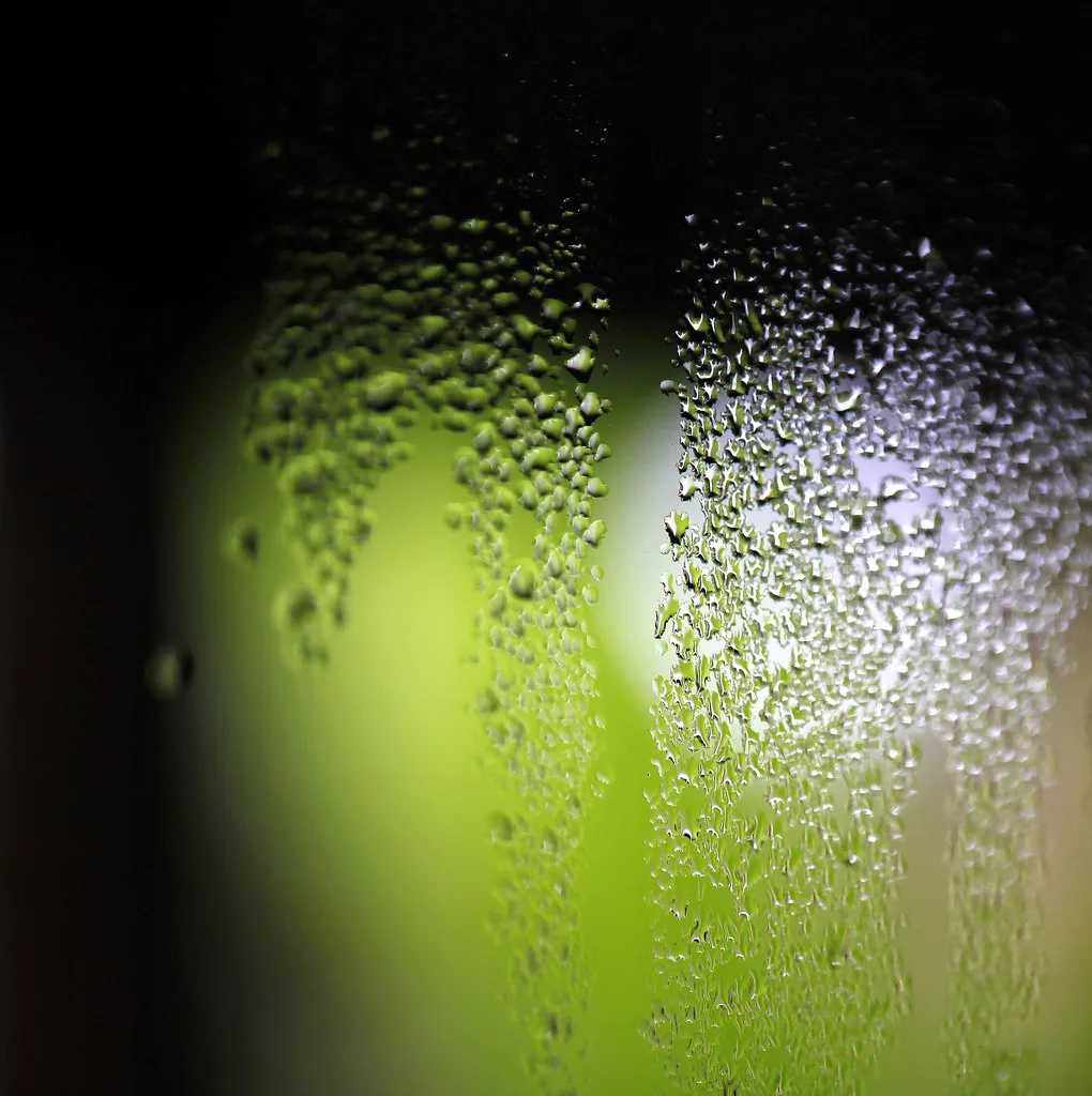 A hand holds a hygrometer beside a fogged window with water droplets and peeling paint.