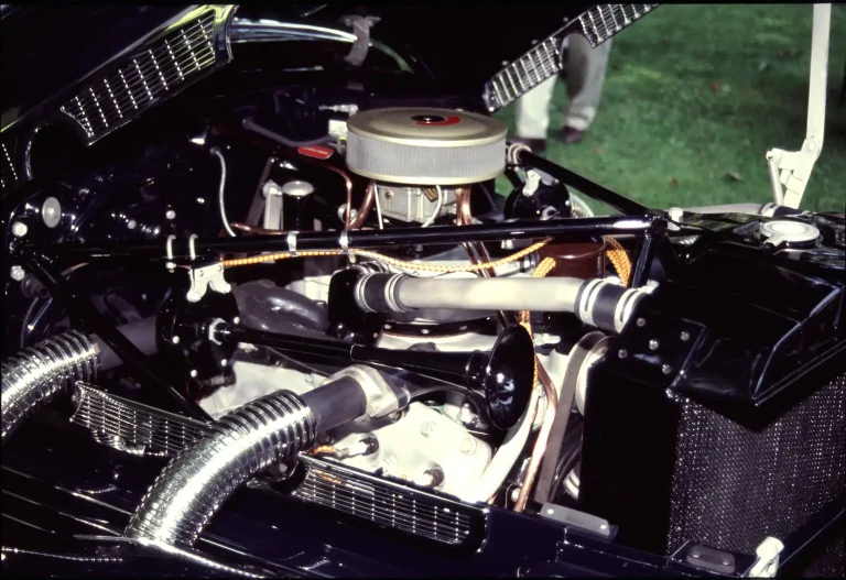 A mechanic tracing a block heater cord path in the open engine bay of a sedan.